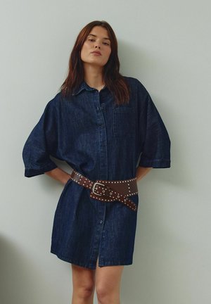 Haljina od trapera tamnoplave boje s prednjim kopčanjem na kopče, širokim rukavima i džepom na prsima, stegnuta oko struka smeđim remenom s zakovicama.