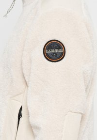 Giacca in fleece colore avorio con tessuto testurizzato, dotata di una toppa circolare nera e arancione sul manica e una tasca con zip laterale.