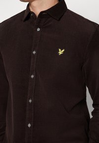 Camisa de pana marrón de manga larga, con cuello tipo botonera, cinco botones y un pequeño logo bordado de un pájaro amarillo en el pecho.