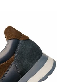 Teniska s tmavě modrým koženým tělem, hnědou semišovou látkou a šedou gumovou podrážkou; má zaoblený tvar a kontrastní švy.