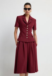 Donna che indossa un blazer corto bordeaux con bottoni perla e una gonna midi plissettata abbinata, accessoriata con occhiali da sole neri a forma di occhio di gatto e orecchini d'oro.
