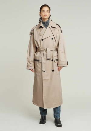 Personne portant un long trench-coat beige croisé avec ceinture, col large et boutons foncés, associé à un jean bleu et des bottines noires.