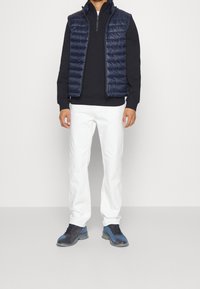 Marinblå quiltad väst över en svart tröja, vita jeans och blågrå sneakers. Västen har dragkedja och hög krage.