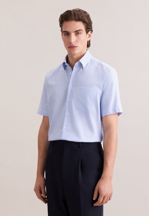 Jonge man draagt een lichtblauw geruit kort shirt dat in een donkerblauwe broek met hoge taille is gestopt, staand tegen een effen achtergrond.