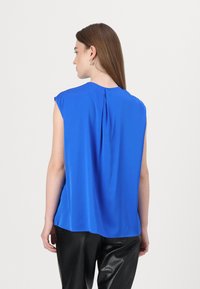 Top senza maniche blu in un tessuto morbido, con una piega sul retro e un colletto arrotondato, abbinato a pantaloni neri testurizzati.
