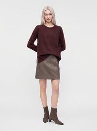 Femme aux longs cheveux blonds portant un pull bordeaux foncé, une jupe en cuir marron et des bottines assorties, debout devant un fond clair.
