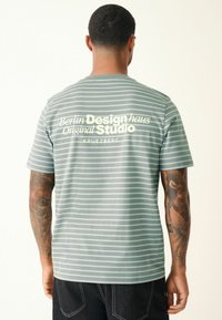 Kurzärmliges, mintgrünes gestreiftes T-Shirt mit weißen Akzenten; auf der Rückseite steht der Text "Berlin Design haus Original Studio"; aus Baumwolle.