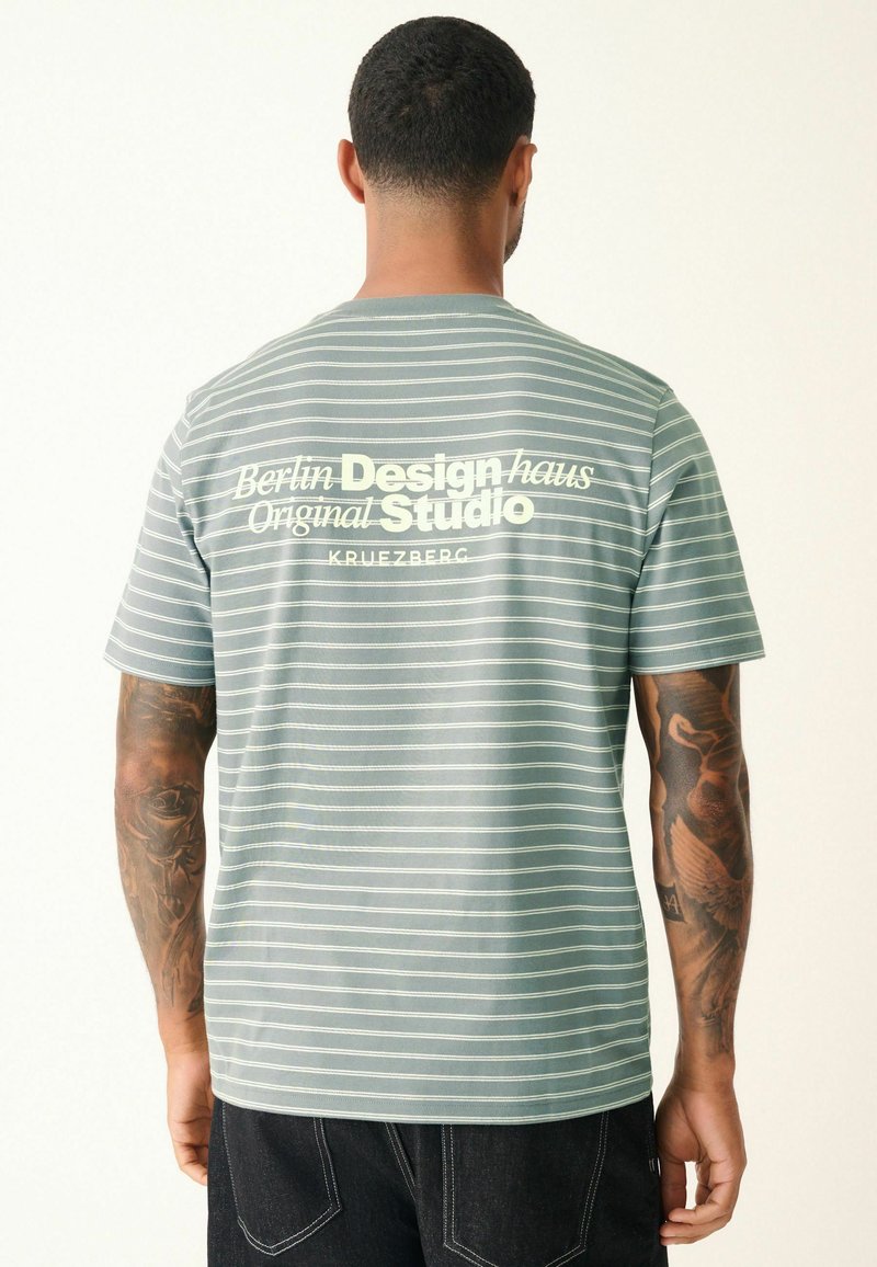 Kurzärmliges, mintgrünes gestreiftes T-Shirt mit weißen Akzenten; auf der Rückseite steht der Text "Berlin Design haus Original Studio"; aus Baumwolle.
