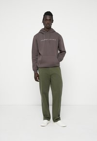 Hanorac maro cu glugă, având logo-ul "TOMMYJEANS", asortat cu pantaloni verzi măslinii și adidași albi, stând pe un fundal simplu.