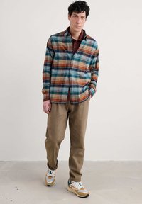 Camicia a quadri con tonalità di blu, arancione e verde, abbinata a pantaloni beige e sneaker multicolori. Tessuto liscio.