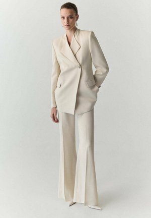 Mujer que lleva una blazer oversize de color crema y pantalones de pierna ancha con zapatos puntiagudos, de pie frente a un fondo claro y liso.