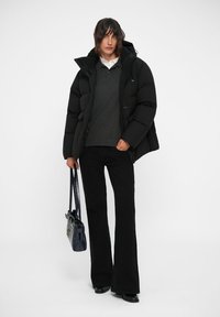 Doudoune noire avec capuche, associée à un pull gris foncé et un pantalon noir évasé. Tenant un sac à main bleu marine, debout sur un fond uni.