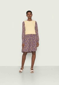 Suéter cropped amarelo claro por cima de um vestido longo de manga comprida, floral roxo, até ao joelho, com um corte descontraído e cintura franzida. Sandálias brancas.
