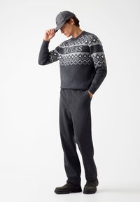 Pull en tricot gris avec des motifs géométriques blancs et logo "GUESS", associé à un pantalon gris foncé et des bottes noires, accessoirisé avec une casquette grise.