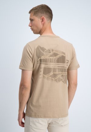 Man die naar links kijkt en een beige T-shirt toont met een architectonisch gebouwontwerp en de tekst "EDGE" op de achterkant.