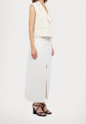 Kobieta w bezrękawnikowym kremowym blezerze, długiej białej spódnicy z przednim rozcięciem i czarnych sandałach na wysokim obcasie ze paskami, stoi bokiem.