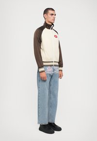 Beige en bruine zip-up jack met een hoge kraag en gestreepte accenten. Model draagt lichtblauwe denim jeans en zwarte schoenen, staande rechtop.