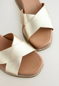 Fémes arany keresztpántos szandálok texturált felülettel, négyzet alakú orral és natúr béléssel. Világosbarna varrással és alacsony sarokkal rendelkeznek.