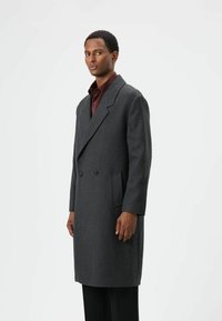 Donkergrijze wollen overjas met een dubbelgeknoopt ontwerp, revers met inkeping en zijzakken, gedragen over een donkerrode blouse. Gladde textuur, nauwsluitende pasvorm.