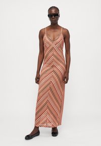 Maxi dress with a chevron pattern in red, orange, and cream; features thin straps and a V-neck; made of lightweight fabric. Black flats worn.