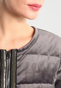 Veste matelassée grise avec une texture douce, dotée d'une fermeture éclair frontale et de bandes décoratives à paillettes le long de l'ouverture.