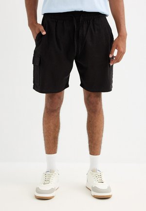 Hombre con pantalones cortos cargo negros con cintura ajustable con cordón, calcetines blancos y zapatillas blancas, de pie sobre un fondo liso.