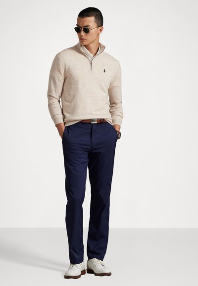 Homme portant un pull beige à zip quart, une chemise blanche, un pantalon bleu marine, une ceinture marron, des chaussures blanches et des lunettes de soleil, se tenant avec les mains dans les poches.