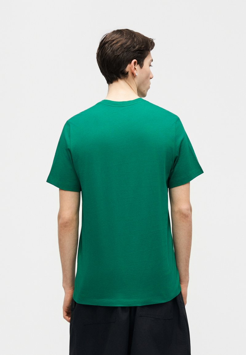 Mann mit kurzen dunklen Haaren, der ein schlichtes grünes T-Shirt mit kurzen Ärmeln trägt, von hinten vor einem schlichten hellen Hintergrund gesehen.