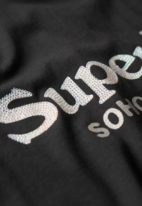 Acercamiento de tela negra con el texto "Sure" decorado con pequeños brillantes y parcialmente impreso "SOHo" debajo.