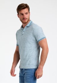 Camisa polo azul claro con una textura de patrón, tapeta de tres botones y cuello y puños a rayas. Combinada con jeans de mezclilla.