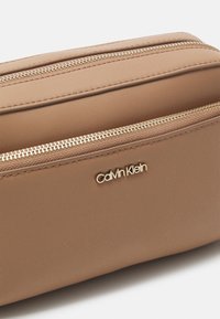 Skórzana kosmetyczka w kolorze brązowym z złotymi detalami, posiadająca dwie zapinane na zamek przegródki oraz subtelnie wytłoczone logo z przodu.