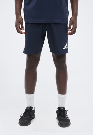 Mand iført mørkeblå atletikshorts med Adidas-logo, hvide crew-sokker og sorte Adidas løbesko, stående mod en ensfarvet baggrund.