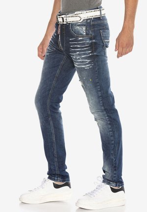 Verblasste blaue Denim-Jeans mit abgenutzten Details und Farbspritzern, im Slim-Fit-Stil, fünf Taschen und kombiniert mit weißen Sneakers.