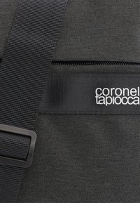 Bolsa de tela negra con material texturizado, una correa ajustable, un bolsillo con cremallera y un detalle de logo blanco en el lateral.