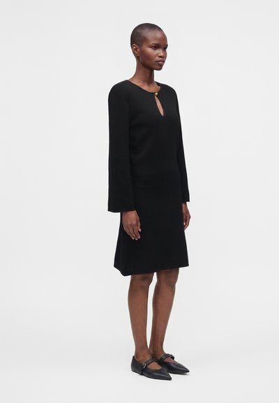 Femme debout portant une robe noire mi-longue à manches longues avec une petite ouverture en forme de goutte d'eau au niveau du col, fermée par un seul bouton.