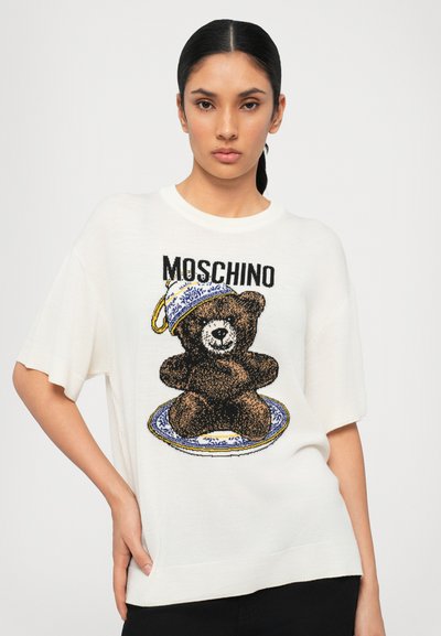 Maglione bianco a maglia con una grande grafica di un orso bruno e un design di piatto blu e bianco. "Moschino" è stampato sopra l'orso. Maniche corte.