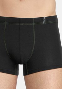 Black boxer shorts made of stretchy fabric, featuring green stitching details, a snug fit, and a logo tag on the waistband.