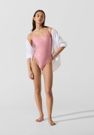 Mladá žena stojí bosá, oblečená v ružové jednodielne plavky a bielu košeľu prehodenú cez ramená na jednoduchom sivom pozadí.