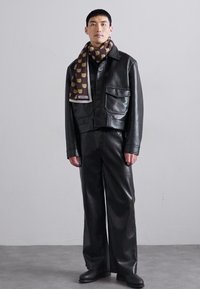 Jachetă scurtă din piele neagră și pantaloni evazați, asortați cu o eșarfă maro cu modele de urși, stând pe un podea gri.
