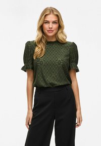Groene blouse met pofmouwen en een gestructureerd patroon, met ruches bij de manchetten, gecombineerd met zwarte high-waisted broeken.