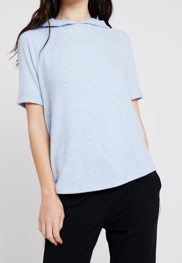 Femme portant un haut à capuche à manches courtes bleu clair et un pantalon noir, debout devant un fond uni.