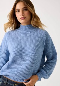 Maglione blu lavorato a maglia con un alto collo a coste, caratterizzato da un design testurizzato e maniche lunghe e morbide. Indossato sopra pantaloni in denim scuro.