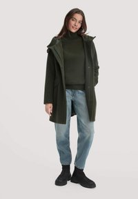 Dunkelgrüner, übergroßer Mantel mit Kapuze, seitlichen Taschen und Druckknopfverschlüssen, kombiniert mit einem passenden Pullover, hellblauen Jeans und schwarzen Stiefeln.