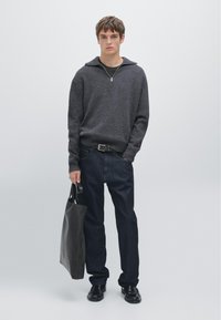 Pull gris côtelé avec un col à fermeture éclair, jean foncé et chaussures en cuir noir. Tenant un sac fourre-tout noir, debout devant un fond neutre.