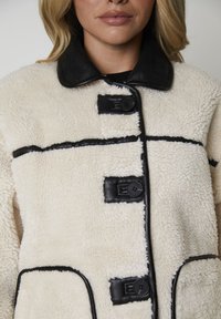 Cremefarvet lammeskindjakke med sort læderkrave og detaljer, der har fire store knapper, kontrasterende syninger og sidelommer.