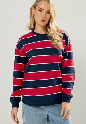 Vrouw met lang golvend blond haar, draag een marineblauw en rood gestreepte trui met lange mouwen en lichtblauwe spijkerbroek, staand tegen een effen achtergrond.