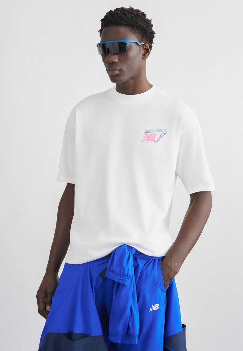 Άνδρας που φοράει λευκό μπλουζάκι New Balance και μπλε αθλητικές σορτς με ταιριαστό μπουφάν δεμένο στη μέση, φοράει μεγάλα μαύρα αθλητικά γυαλιά ηλίου.