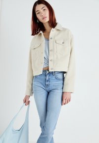 Casaco cropped creme com um acabamento texturizado, dois bolsos no peito, botões prateados; combinado com jeans azul claro e segurando uma mala azul clara.