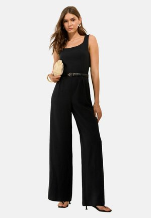 Mujer con un mono negro sin mangas con cinturón, sosteniendo un bolso de mano beige tejido, lleva pendientes de aro y sandalias de tacón sobre fondo blanco.