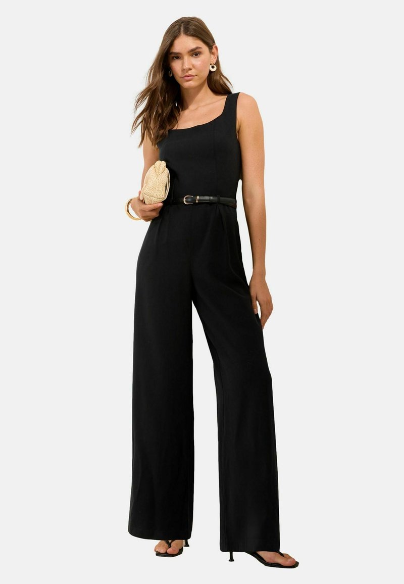 Kobieta w czarnym kombinezonie bez rękawów z paskiem, trzymająca beżową plecioną kopertówkę, nosząca kolczyki-koła oraz sandały na obcasie na białym tle.